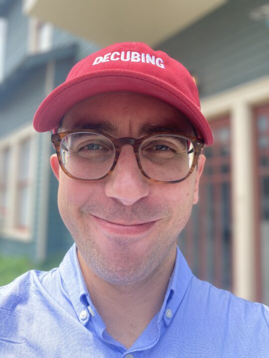 Headshot of Blake Bertuccelli-Booth, a smiling male with glasses and a red hat that reads "Decubing".